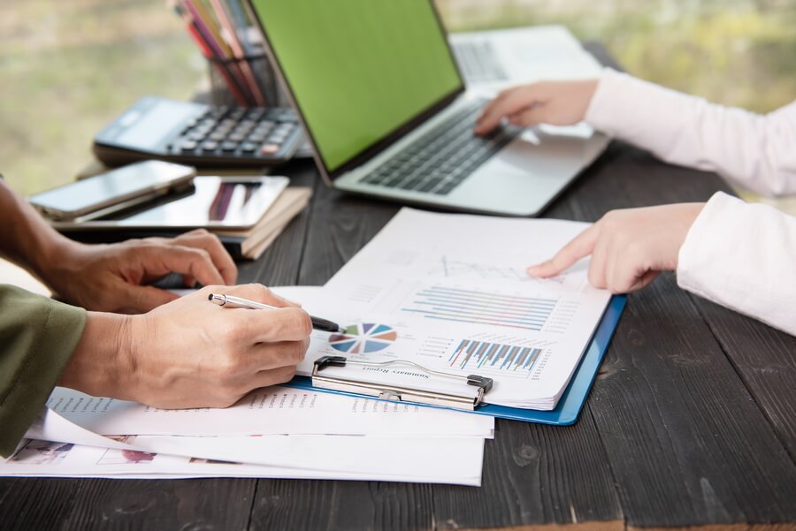 Two colleagues at a desk working through a printed report on a clipboard, one pointing at a row of figures while the other annotates a page beside a calculator and a laptop - the annual assembly of ACA employer reporting before the furnish and file deadlines
