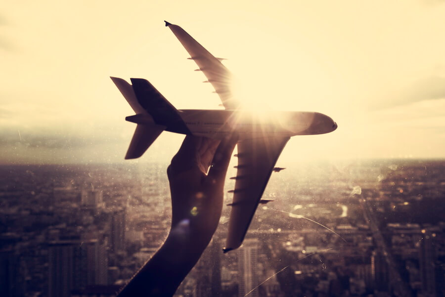 A hand holding a model airliner silhouetted against the sun above a hazy city skyline