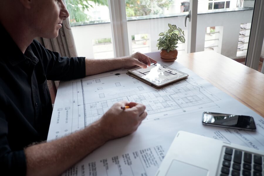 A design professional marking up a printed floor plan at a desk beside a tablet showing the same drawing, while the practice tracks every license, PDH deadline, and firm registration