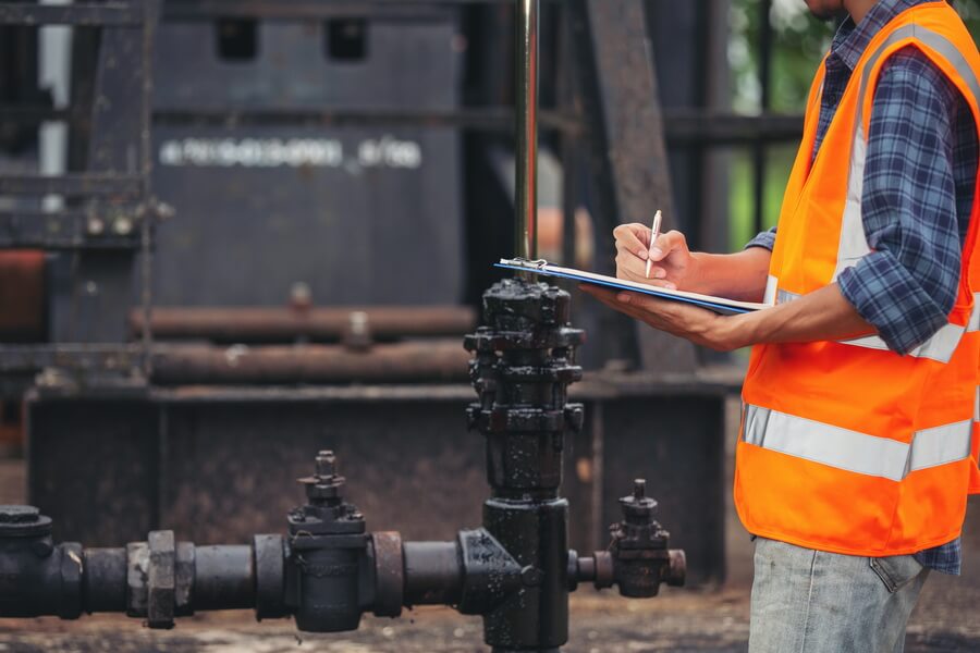 A worker in a hi-vis vest writing on a clipboard beside outdoor cast-iron pipework and valves - recording a field test on site, which is only the first half of a backflow test that also has to be reported to the water utility