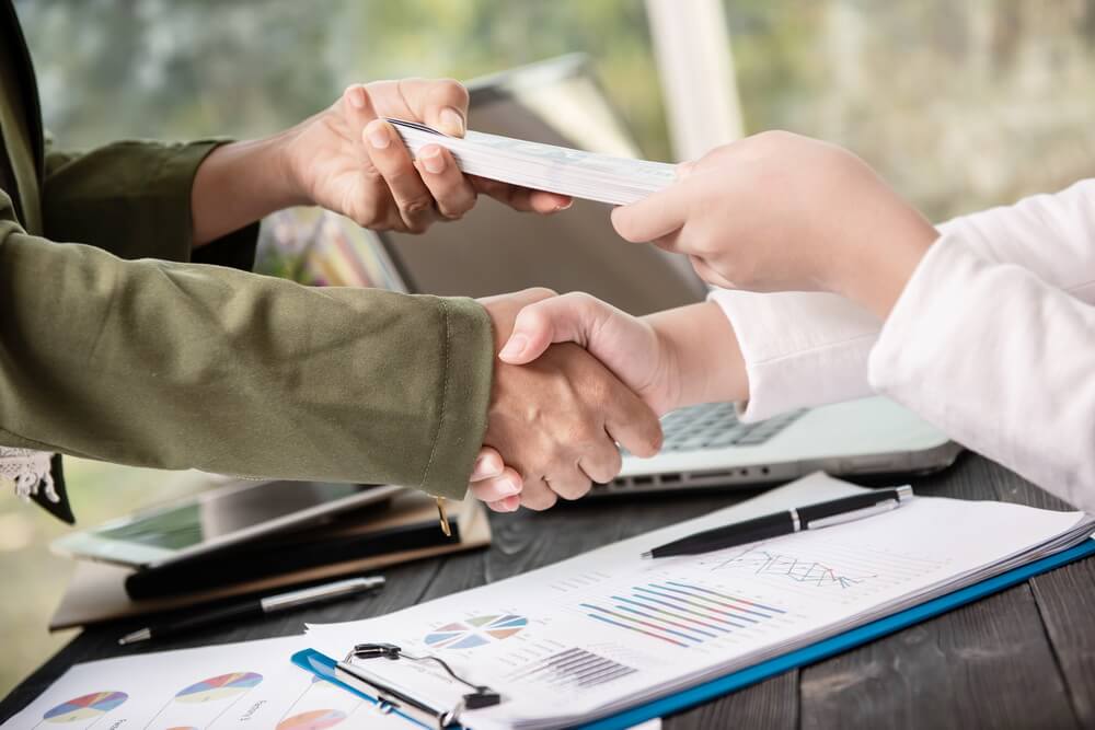 Two parties shaking hands across a desk as a document changes hands beside financial charts — the moment a bank guarantee is put in place and then forgotten