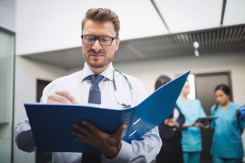 A physician in a white coat and stethoscope reviewing a file in a hospital corridor, with clinical staff behind — keeping every provider's board certification and MOC requirements current