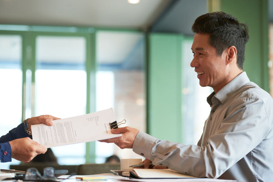 A person handing a clipped set of documents across a desk to a colleague in an office
