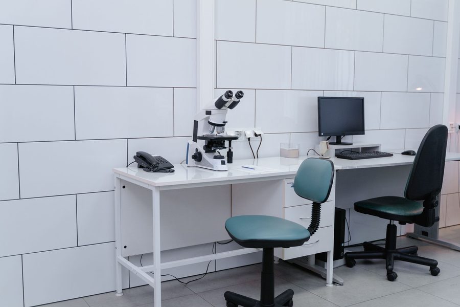 Clinical lab bench with white microscope, telephone, and papers beside a computer workstation and chairs against tiled walls