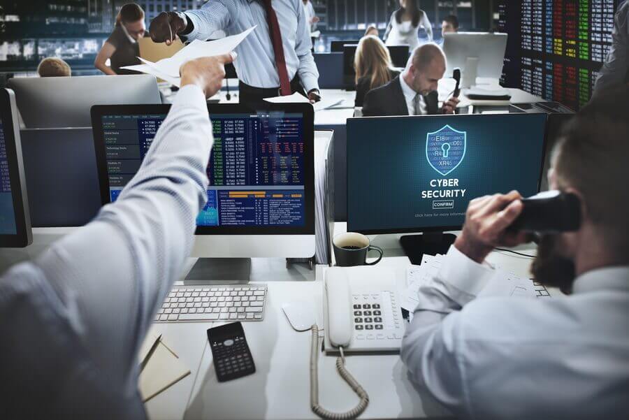 Staff working at desks in a busy contractor office, one on a desk phone amid paperwork, with a cyber security shield graphic on one of the monitors