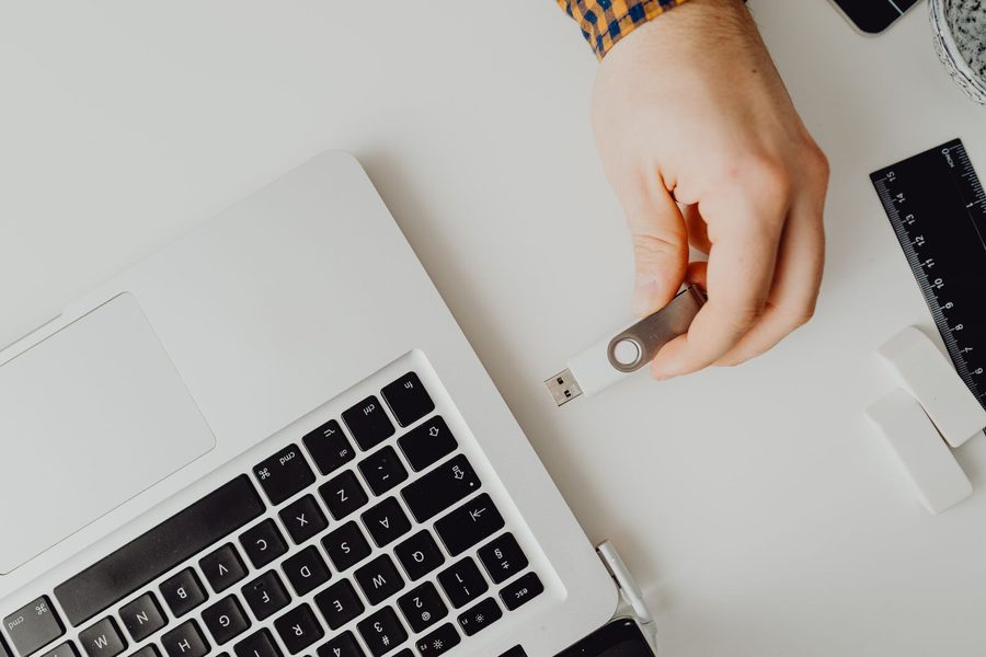 Overhead view of a hand holding a USB drive beside an open laptop on a clean white office desk