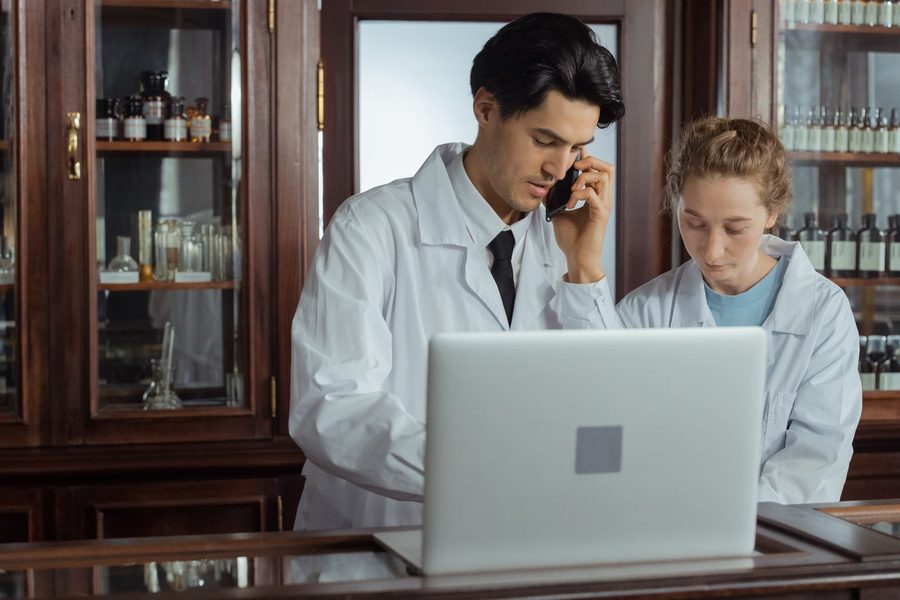 Two pharmacists in white coats at a pharmacy counter, reviewing a laptop while one takes a call