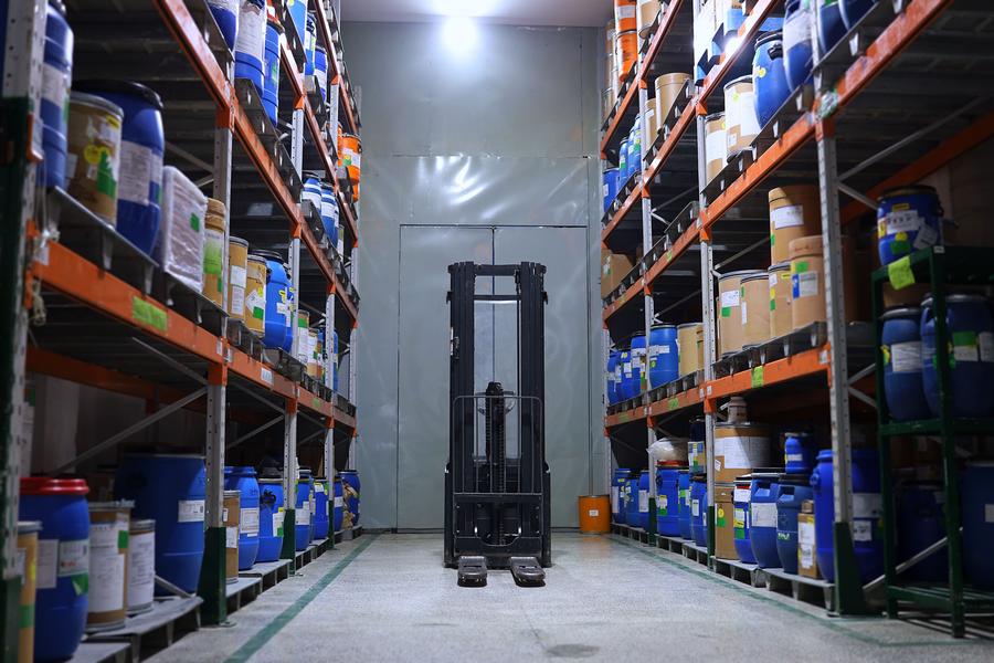 Aisle of a chemical distribution warehouse with labeled drums and fiber containers stored on orange pallet racking, with a forklift at the far end