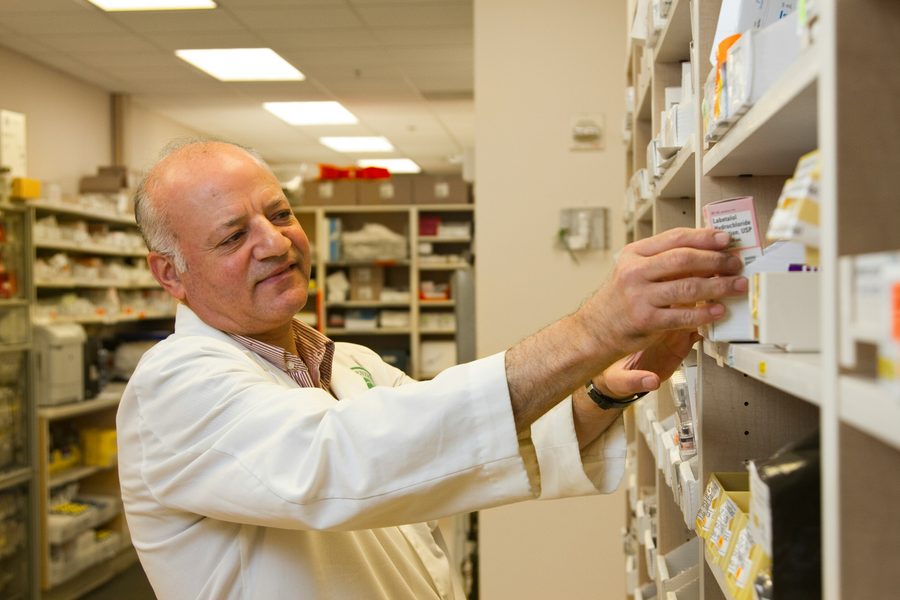Pharmacist in a white coat pulling a medication box from dispensary shelving, with rows of stocked pharmacy shelves behind him