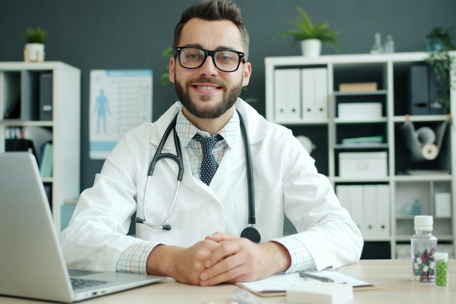 Smiling doctor in a white coat and stethoscope seated at a desk beside an open laptop, ready for a telehealth video visit