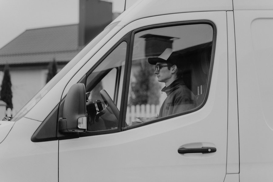 Black and white side view of a fleet driver in glasses and a cap seated at the wheel of a white cargo van on a residential street