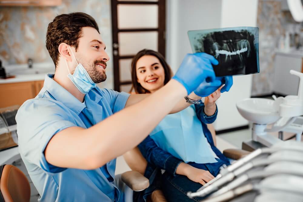 A licensed dentist reviewing a dental X-ray with a patient in the chair — clinical care that requires current dental licenses and permits