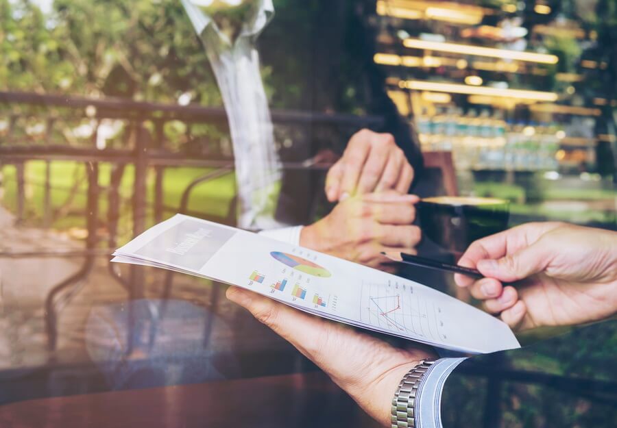 Two colleagues at a table reviewing a printed summary report of charts, one gesturing with a pen - for EEO-1 reporting, assembling the numbers is the routine part and the filing deadline is the part nobody can look up