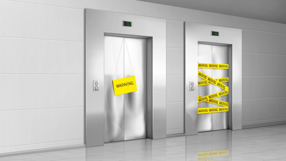 Two elevator doors in a lobby closed off with a warning sign and hazard tape — cars taken out of service after a compliance lapse