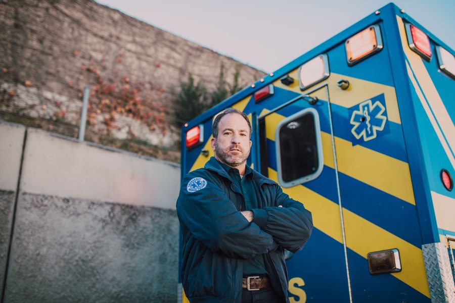 EMT in uniform standing beside a blue and yellow ambulance outdoors