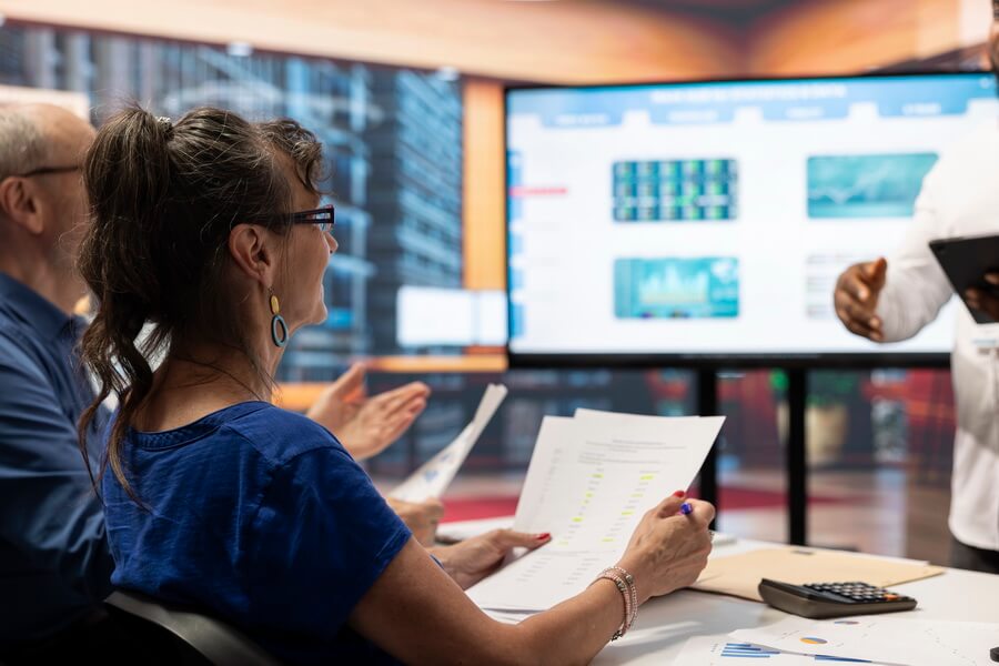Three colleagues around a conference table reviewing a printed report line by line against figures on a wall display, one of them highlighting rows with a pen - the desk-bound work of assembling an annual chemical inventory report before its March 1 deadline