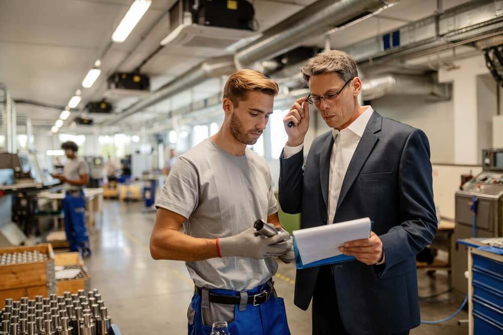 A technician holding a machine component while a manager checks the paperwork on a factory floor — establishing whether an asset is still under warranty before a repair is authorized