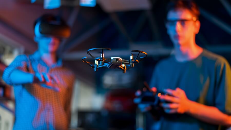 A small quadcopter with propeller guards hovering indoors under blue and orange light between two operators - one wearing a headset, one holding the controller - a scene that looks identical whether the flight is recreational or commercial