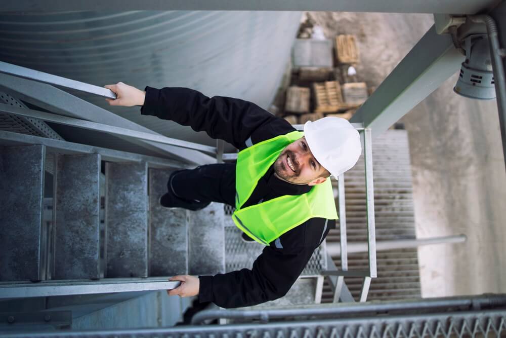 A hard-hatted worker in a hi-vis vest climbing a steel stairway at height on an industrial structure — the working-at-height work that fall protection training and equipment keep safe