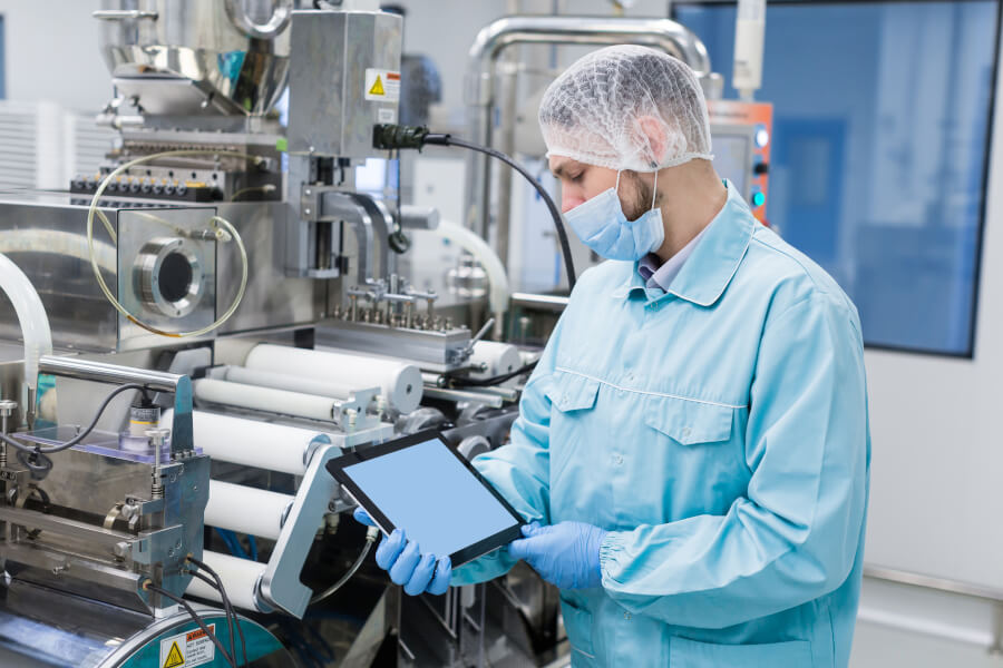 A technician in a light blue cleanroom gown, hairnet, face mask and blue gloves holding a tablet beside stainless steel production and packaging machinery in a clean manufacturing hall