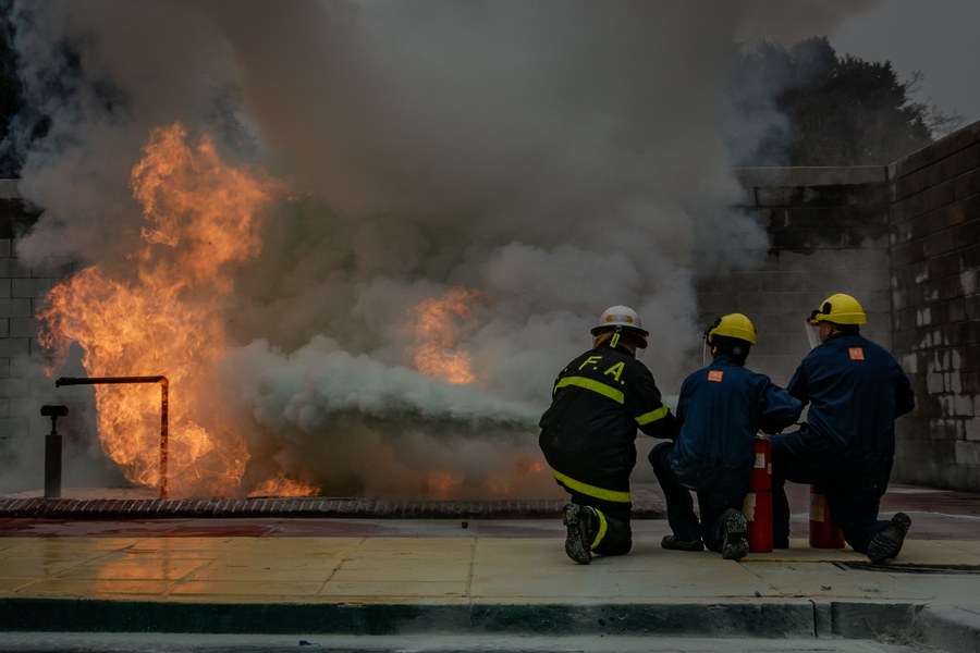 Trainees in hard hats and coveralls kneel beside red fire extinguishers and spray a controlled fire during a hands-on fire safety training session