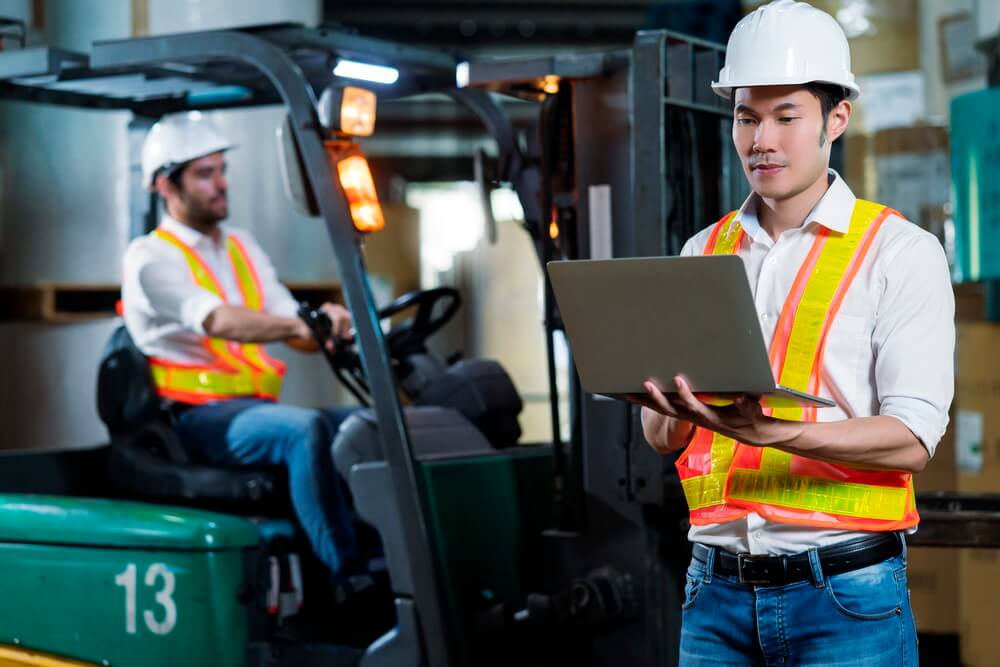 A warehouse supervisor in a hard hat and hi-vis vest checking records on a laptop beside a forklift operator seated on a lift — keeping every operator's certification current on the floor