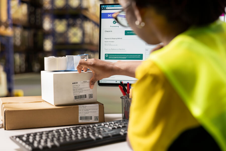 Warehouse operative checking labelled cartons against an on-screen tracking view, representing imported food moving through entry while the supplier verification behind it is tracked separately