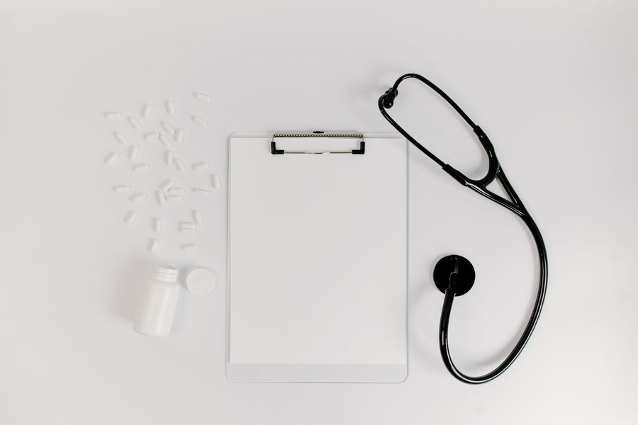 Blank health benefits form on a clipboard beside a black stethoscope and a pill bottle on a white desk