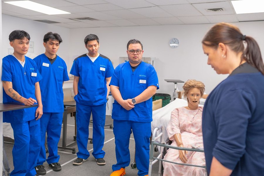 Clinic staff in blue scrubs listen to an instructor during a hands-on training session in a skills classroom