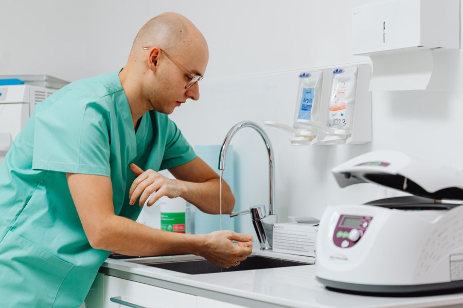 Dental clinician in green scrubs washing his hands at a clinic sink with wall-mounted soap dispensers - hand hygiene for infection control training