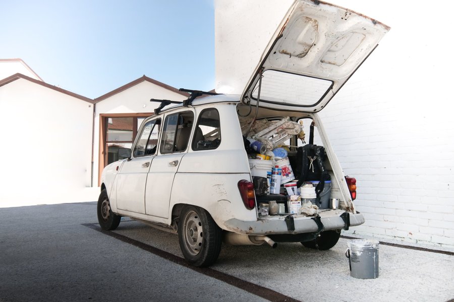 Rear hatch of a well-worn white work car open to show painting supplies, buckets and a stepladder packed in the cargo area, with a paint pail on the ground