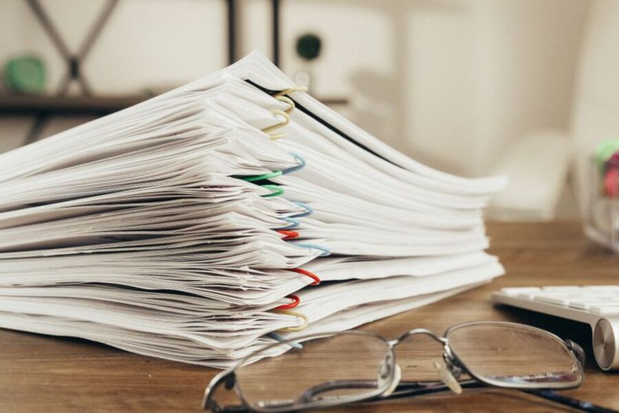 Tall stack of paper documents held with coloured paper clips on a wooden desk beside reading glasses
