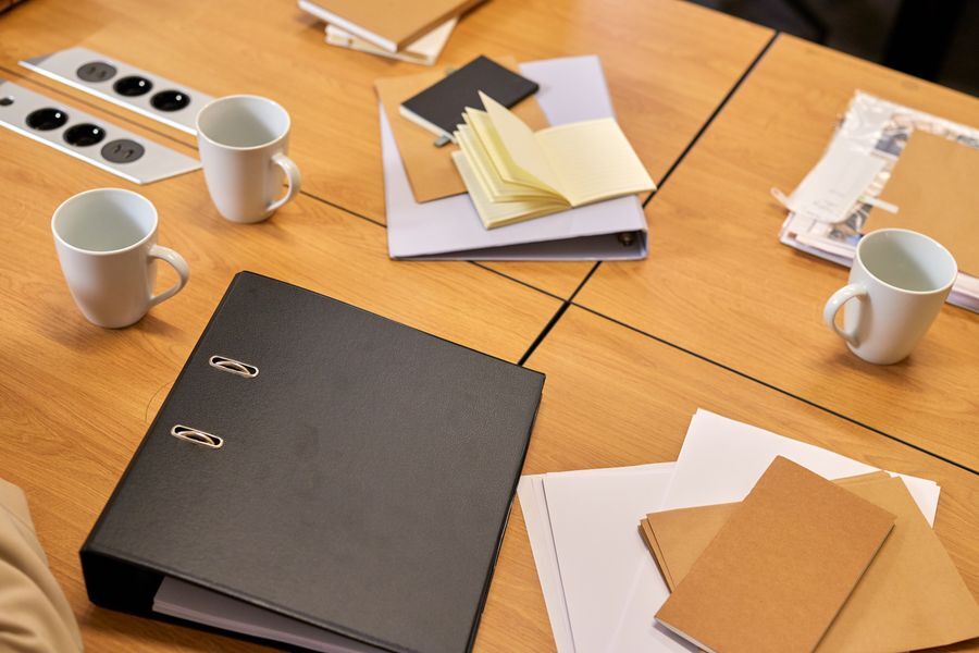 Black ring binder, notebooks and paper folders on a wooden meeting table beside coffee mugs