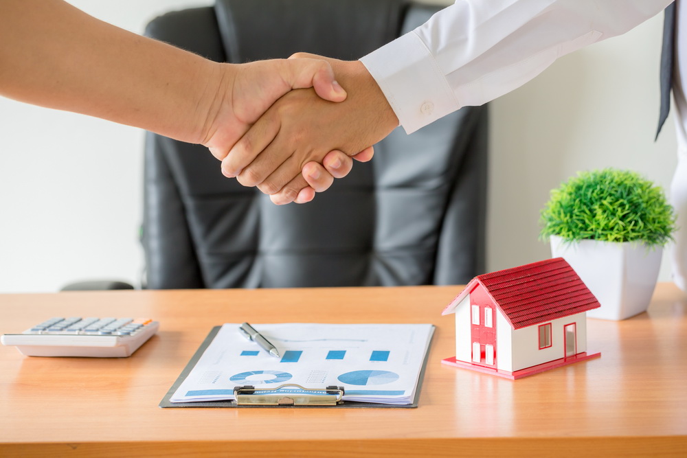 Two people shaking hands over a desk with a model house and signed paperwork — closing a lease agreement before its renewal and notice deadlines