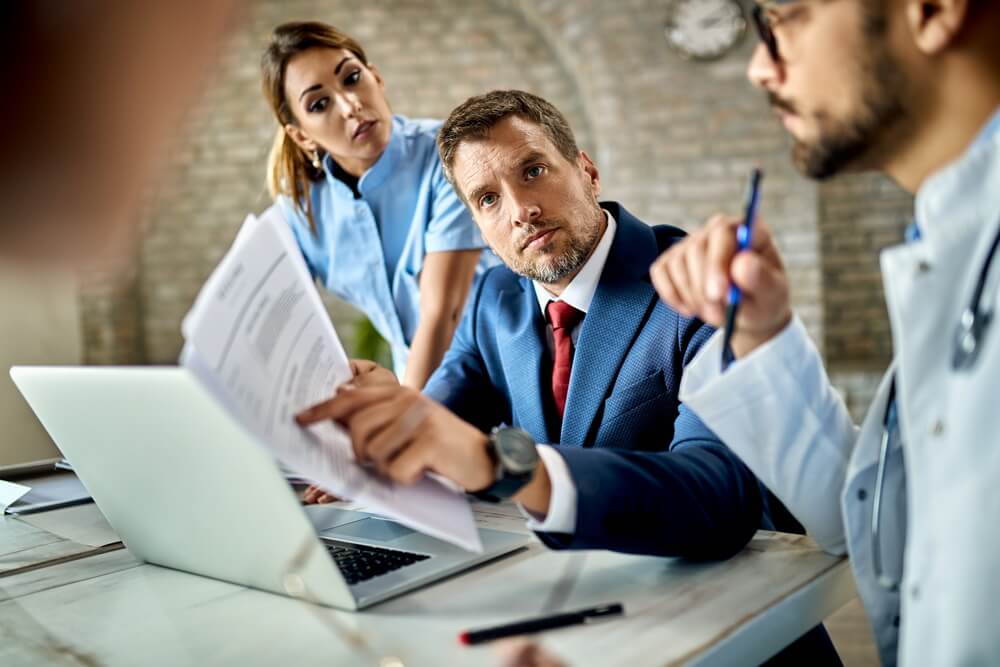 An insurance advisor in a suit reviewing coverage paperwork with a doctor and nurse at a clinic table — keeping every provider's malpractice coverage current