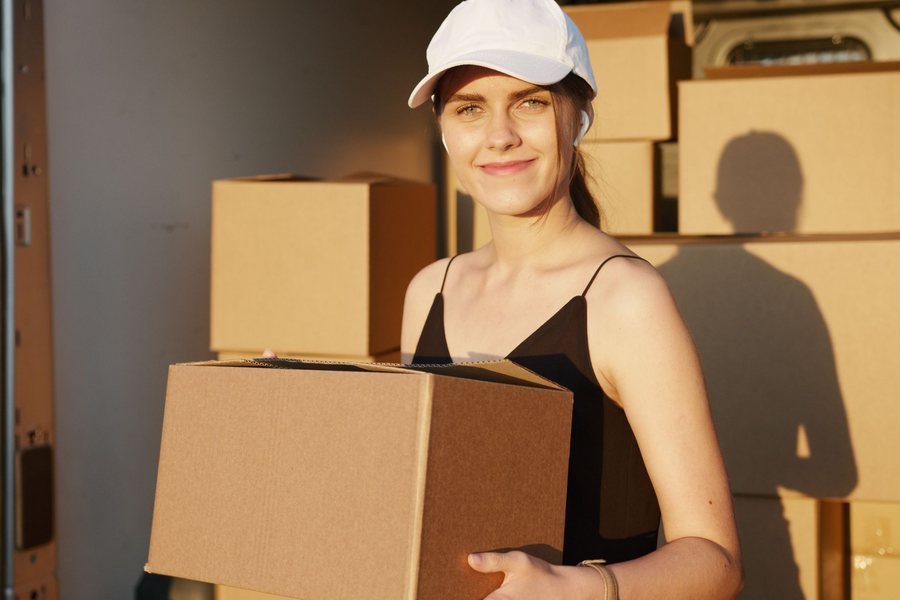 Worker in a white cap holding a cardboard box close to her body at waist height beside a loaded delivery van