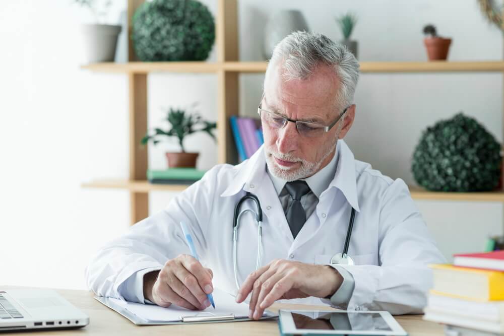 A physician in a white coat and stethoscope reviewing and signing documents at a desk — keeping every state medical license, DEA, and CME current