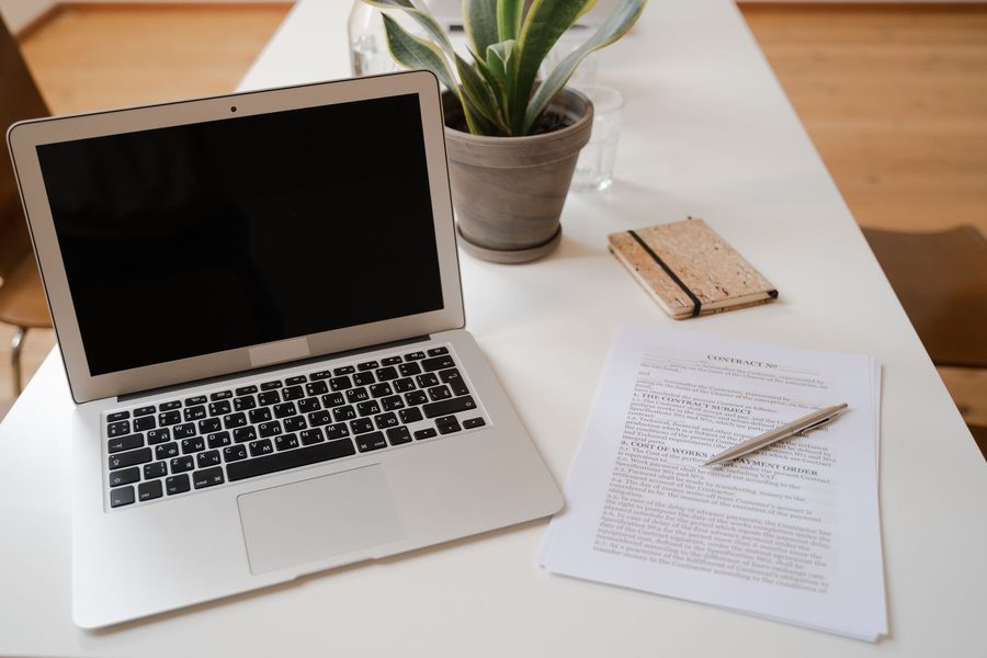 Open laptop beside a contract, pen, notebook, and plant on a white desk