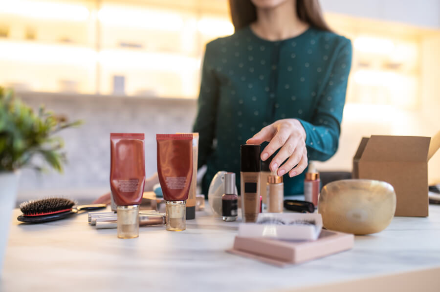 A woman reaching for a foundation bottle among cosmetic tubes, serums and a compact arranged on a marble table beside a shipping box