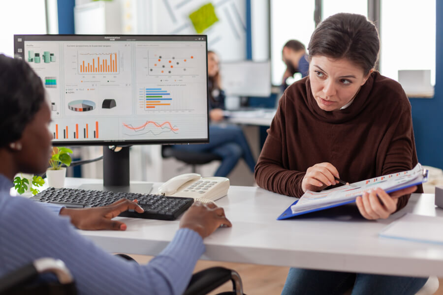 Two colleagues at a shared desk in an open-plan office, one at a keyboard in front of a monitor of charts and the other checking a printed report in a ring binder - the two separate records an employer's new hire reporting deadline can end up being calculated from