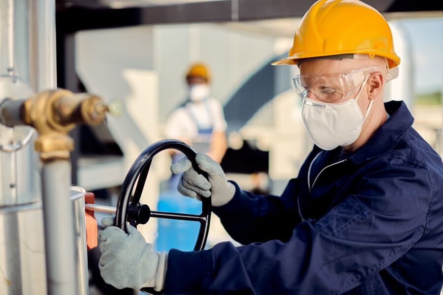 A plant worker in a hard hat, safety goggles, mask and gloves turning a valve wheel on industrial pipework - a year of work like this is what the OSHA Form 300A annual injury summary reports on, before it has to be certified, posted at the site, and submitted to OSHA