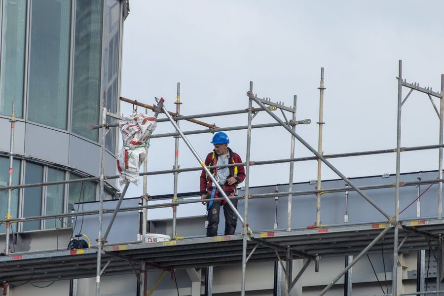 Worker in a hard hat and safety harness standing on a metal scaffold platform beside a glass-fronted building