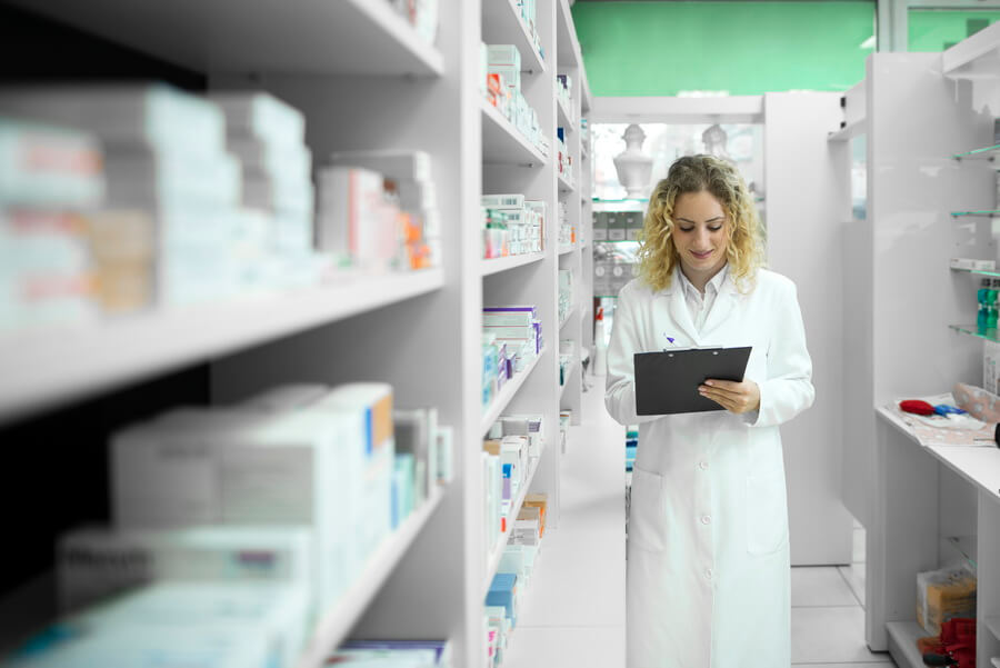 A pharmacist in a white coat checking stock against a clipboard in the aisle between a dispensary's shelves