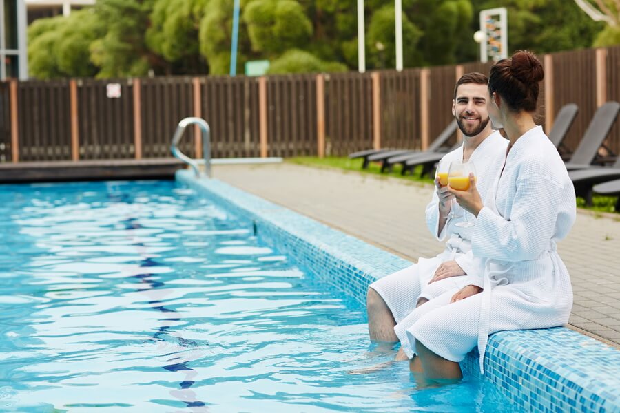 Two guests in white robes sitting on the edge of a hotel pool with their feet in the water