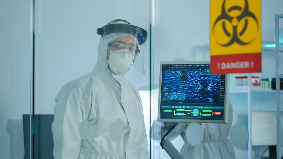A technician in a full protective coverall and face shield working behind a glass partition in a controlled area, beside a monitor showing scan imagery and a hazard warning sign
