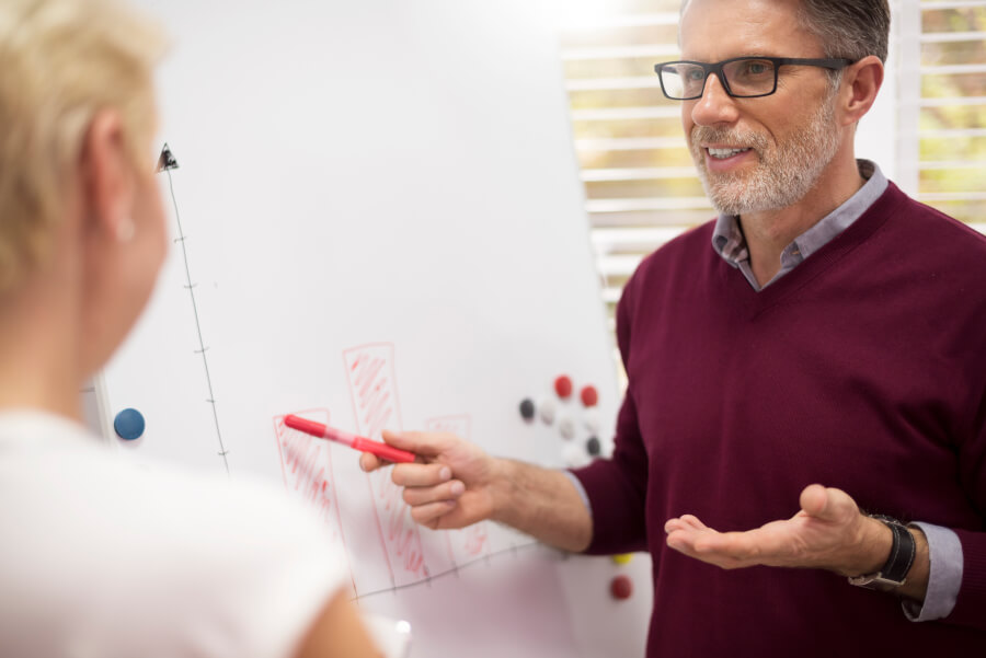 A man in a maroon sweater standing at a whiteboard, gesturing with a red marker toward a hand-drawn bar chart while explaining it to a colleague seated in the foreground of a bright office