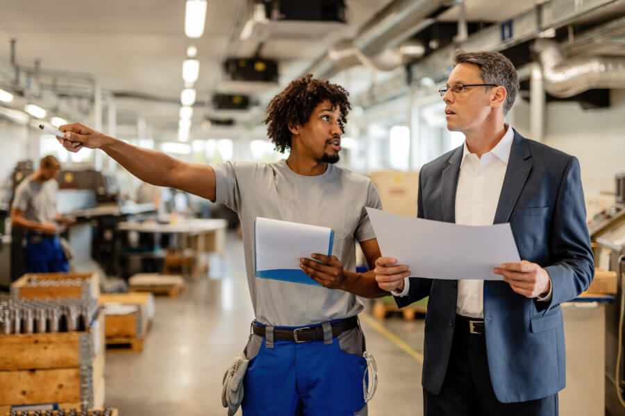 A business owner in workwear showing a visitor holding documents around a small manufacturing floor - the kind of site visit a DBE certifier may make unannounced to review continued eligibility