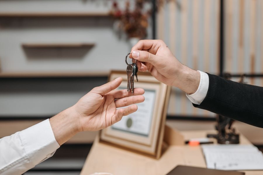 Two hands passing a set of keys across a desk, with a framed certificate and papers behind