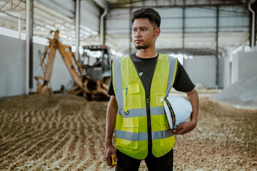 Construction worker in a high-visibility vest holding a white hard hat inside a large site shed with an excavator behind