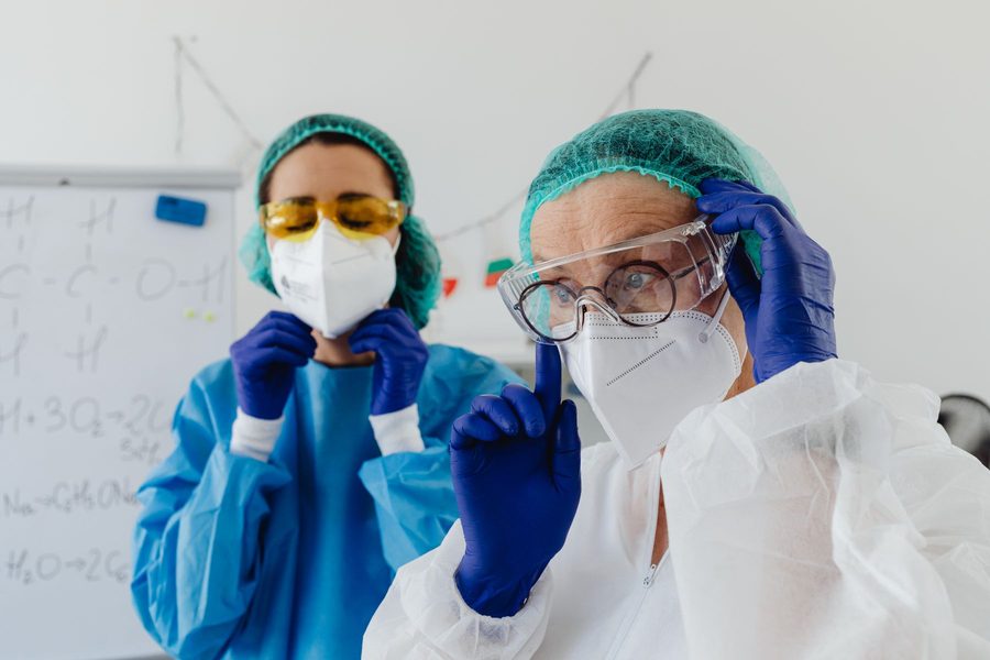 Two lab workers in gowns, gloves and respirator masks putting on safety goggles before handling chemicals